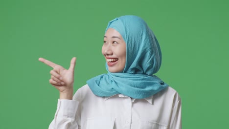 close up of asian muslim woman smiling and pointing to side while standing in the green screen background studio