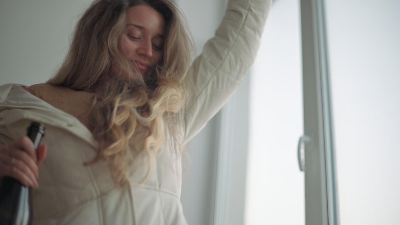 A woman with long blonde hair joyfully dancing while holding a bottle near a bright window
