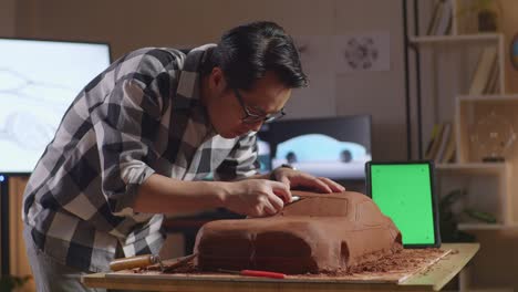 asian man automotive designer with green screen tablet using rake or wire to smooth out the surface and create details in the sculpture of car clay in the studio