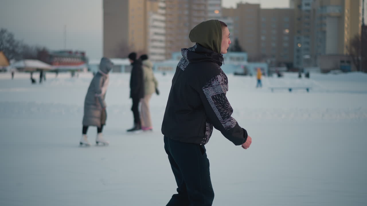 side view of student in patterned winter jacket gliding on icy surface with other skater nearby, snowy urban rink under pale winter sky, smooth ice reflecting soft light for seasonal leisure vibe