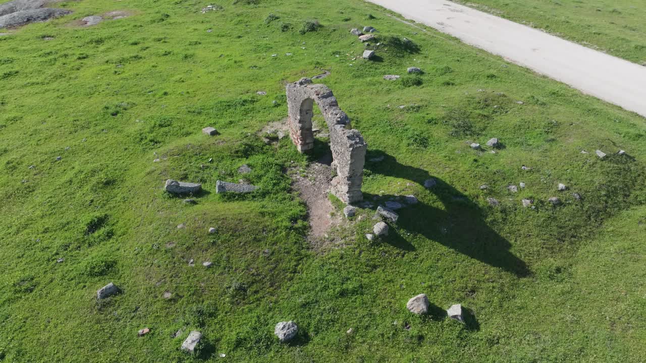 Orbital drone flight around the central stone arch of the Puerta de Arenas. Scattered stones and rectangular mounds beneath the earth reveal the buried remains of its ancient structure.