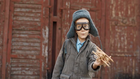 niño pelirrojo sonriente con sombrero y gafas jugando con un avión de juguete de madera y soñando con ser aviador