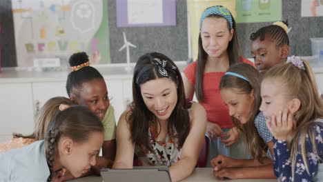 happy diverse female teacher with schoolgirls using tablet in classroom at elementary school