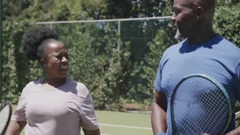 Happy-senior-african-american-couple-walking-with-tennis-rackets-and-talking-at-tennis-court