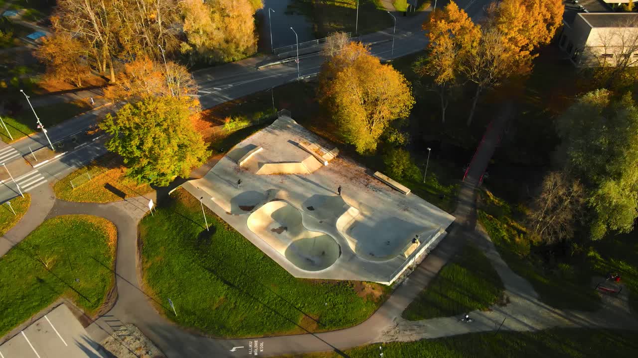 Aerial drone video orbiting and flying around a large gray colored concrete or cement skatepark in a small town grassy green park with autumn trees around with fallen golden yellow leaves, sunny day
