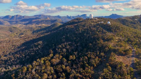 drone footage glides toward a mountaintop observatory complex in warrumbungle national park, revealing telescopes amid forested hills under bright daylight