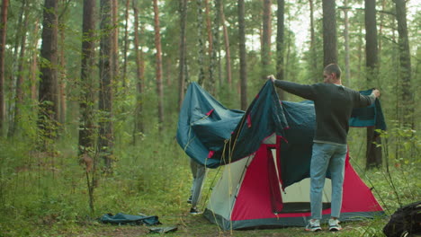 friends work together to cover tent with large fabric sheet in peaceful forest campsite, standing among tall trees and greenery while adjusting material to shield shelter during outdoor setup