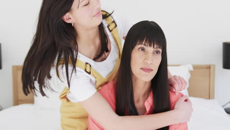 Happy-biracial-mother-and-adult-daughter-sitting-embracing-and-kissing-on-bed-at-home,-slow-motion