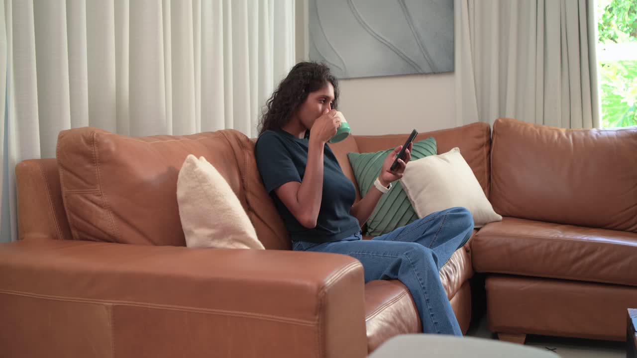Donna afroamericana che legge al telefono, sorride, sorseggia da una tazza e si rilassa sul divano guardando fuori dalla finestra.