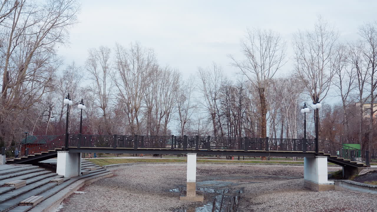 Elevated pedestrian bridge with black railings and white lampposts stretches over dry stony stream in public park, surrounded by leafless autumn trees and people walking in background