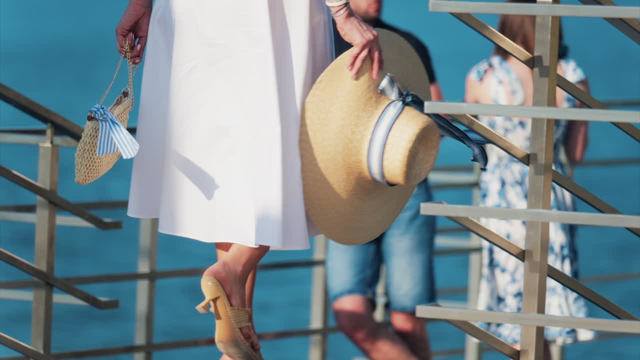 Woman in a white dress, holding a wide hat with a ribbon walking on a scenic terrace with a view of a calm body of water