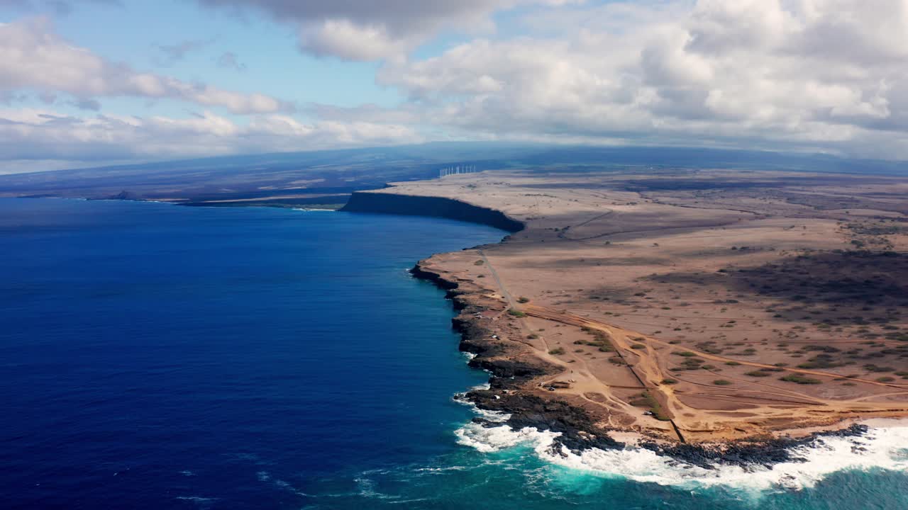 From above, steep lava cliffs meet restless surf along a barren coastal plain, where narrow dirt tracks and wind-shaped terrain stretch into the distance beneath a sky veiled in layered clouds