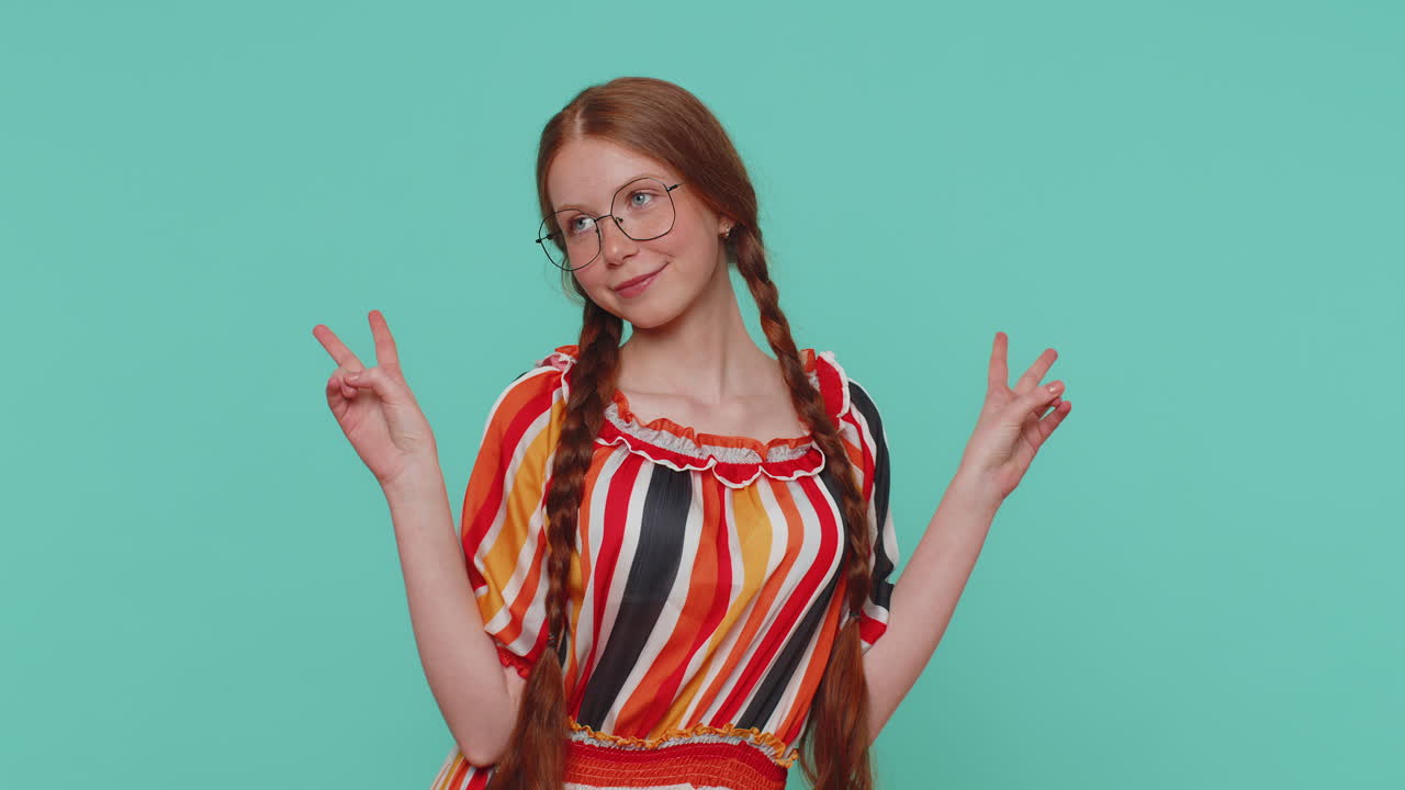 은 머리의 사랑스러운 소녀는 승리의 표시를 보여주고, 성공과 승리를 희망하며, 평화의 몸짓을 하고, 미소 짓고 있습니다.