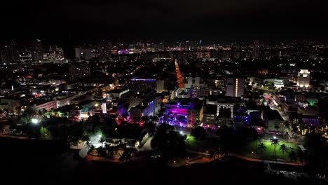 Sugerentes-Imágenes-Aéreas-Nocturnas-De-Drones-De-Miami,-EE.UU.:-Lento-Movimiento-Hacia-Atrás,-Desde-La-Ciudad-Con-Edificios,-Luces-De-Colores,-Carreteras,-Hasta-La-Orilla-Del-Mar-Y-La-Oscuridad-Del-Mar