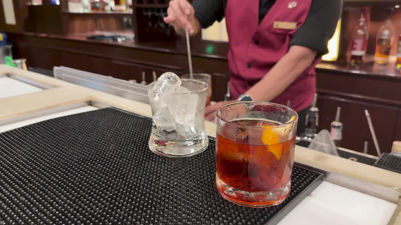 Bartender in red vest stirs cocktail with ice, warm lighting, shallow depth, steady camera