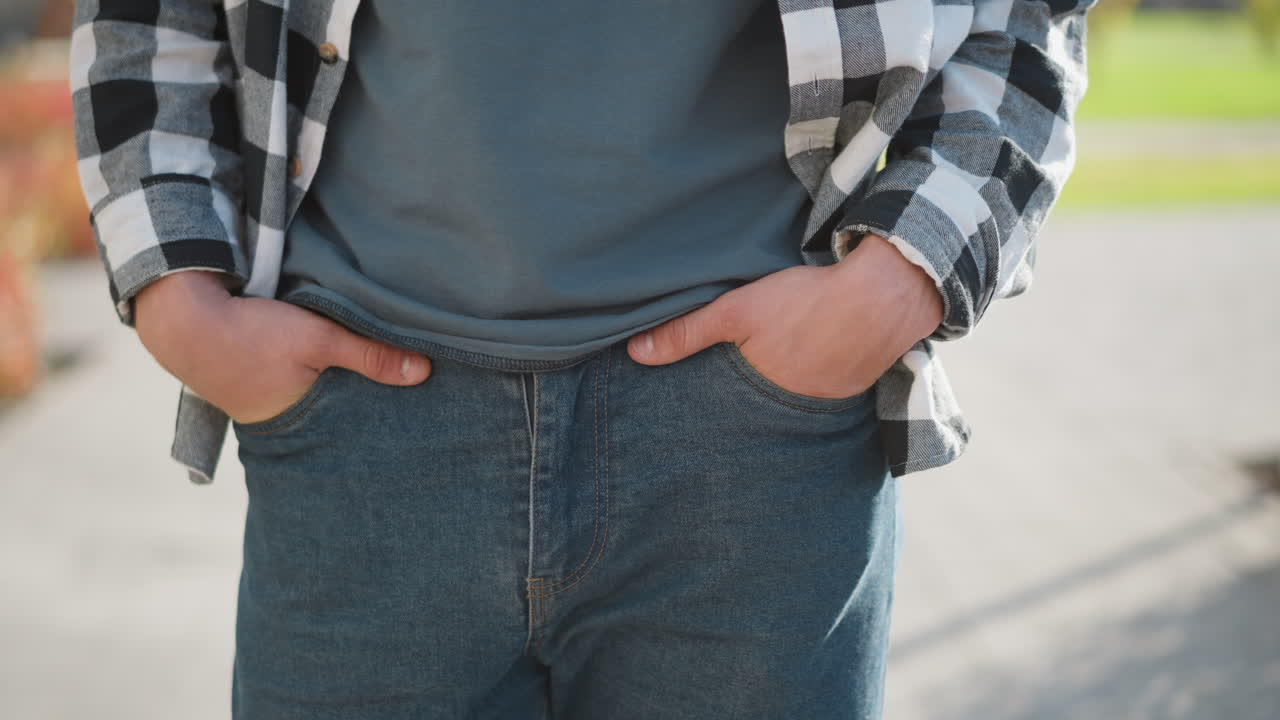 Close up of student strolling slowly with both hands in jean pockets along sunlit paved pathway with soft blur of greenery and red shrubs in background capturing relaxed casual stride