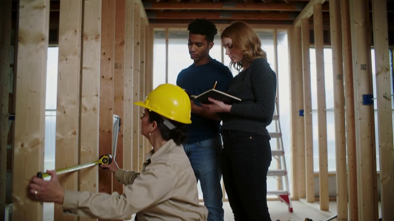 Measuring kneeling worker in coverall checking studs on site for inspection tape hardhat notes