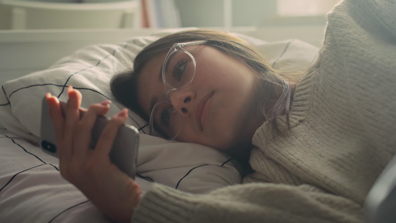 Nærbillede af en hvid teenagepige, der browser på sin telefon, mens hun ligger på siden i sengen.