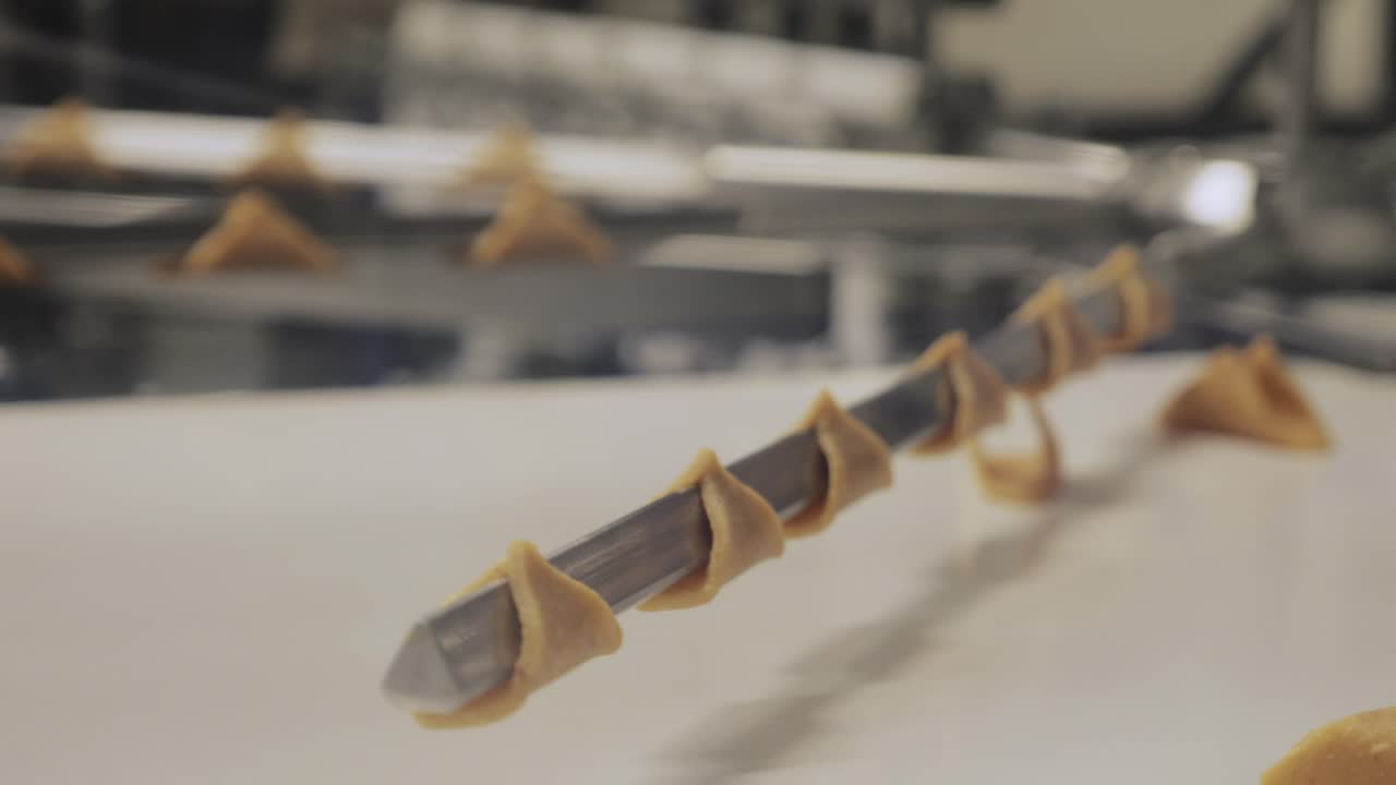 Close-up shot of small triangular dough pieces wrapped around a metal rod on a moving conveyor in a factory setting