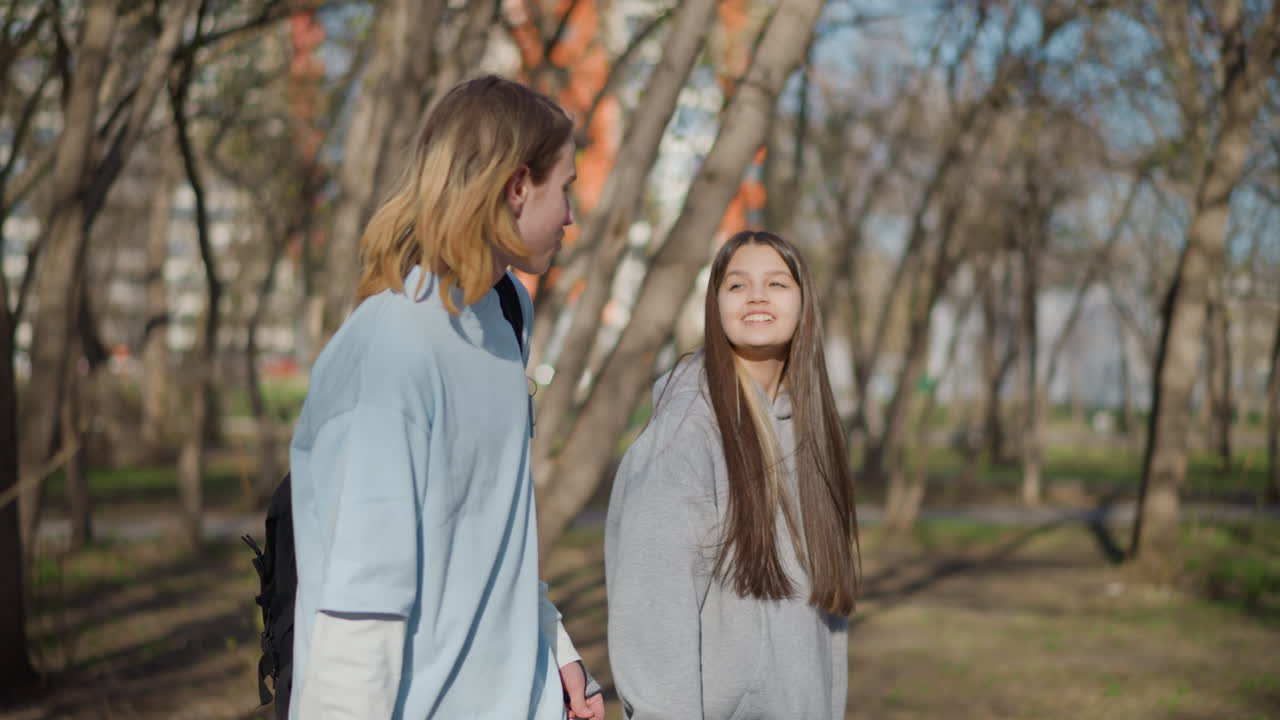 공원 산책로에서 두 젊은 여성이 채팅하고 있고, 편안한 캠퍼스 학생들은 나무 아래 멈춰 서서 미소를 지으며 이야기를 나누고 웃고 있어요. 데님과 후드티를 입고, 부드러운 햇빛이 앙상한 나뭇가지를 통해 비치고 있으며, 도시 배경이 보입니다.