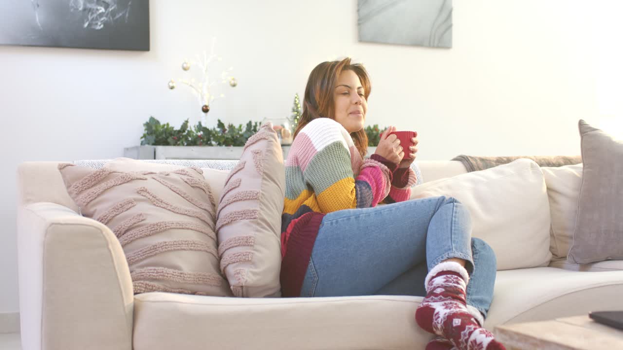 Donna ispanica di mezza età con un maglione a righe sul divano che prende una tazza rossa, inalando il vapore e sorseggiando per scaldarsi.