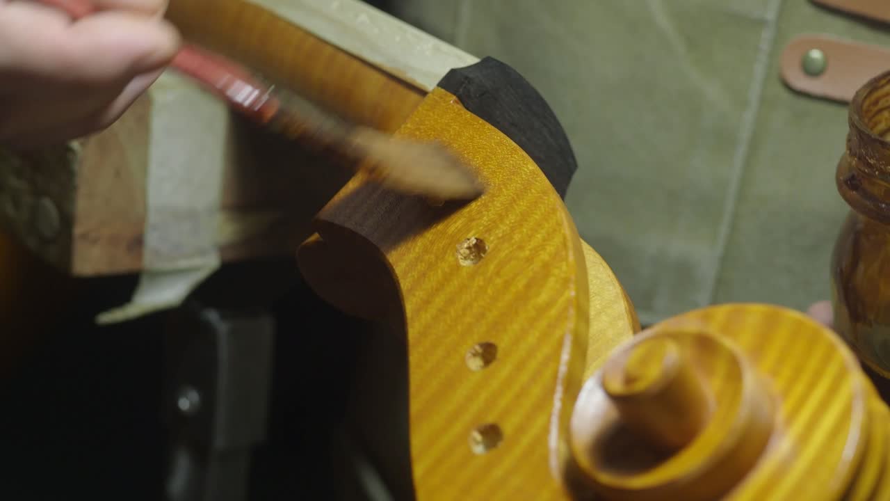 Liutaio al lavoro su cavigliera e riccio di violoncello, applicando con un pennello una vernice naturale fatta in casa in un laboratorio scarsamente illuminato, mettendo in mostra l'artigianato e la precisione coinvolti nella fabbricazione di strumenti musicali