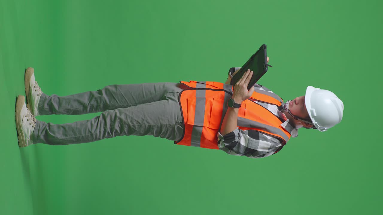 Full Body Side View Of Asian Male Engineer With Safety Helmet Taking Note On The Tablet And Looking Around While Standing In The Green Screen Background Studio
