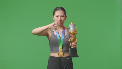 mujer asiática con una medalla de oro y un trofeo señalando y mostrando los pulgares hacia abajo gesto para ridiculizar al perdedor en el fondo de la pantalla verde en el estudio