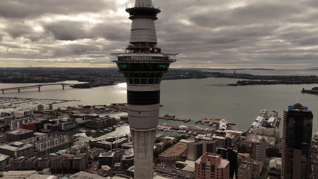 Famous sky tower in Auckland city during cloudy sunset. Aerial backwards wide shot. Harbor bridge in distance, New Zealand