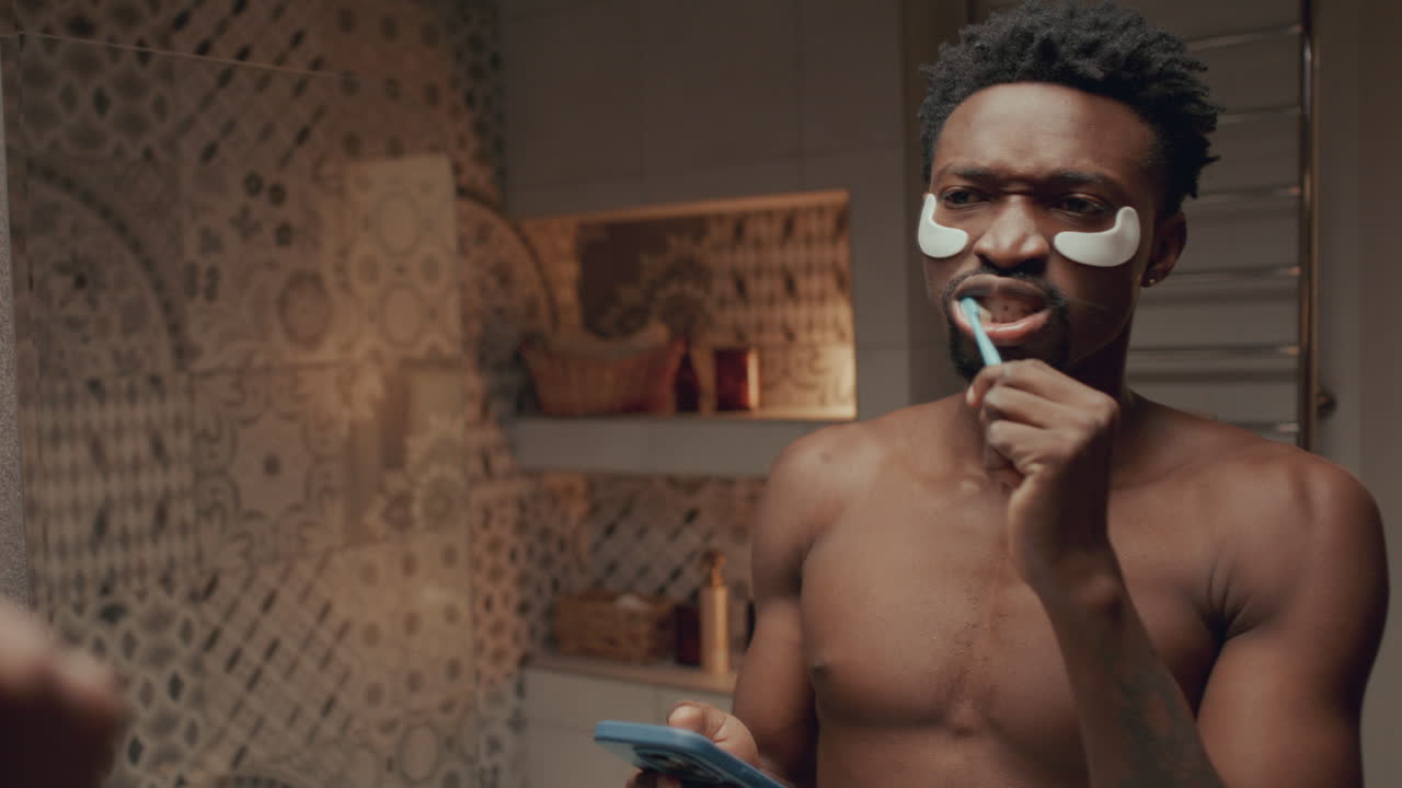 joven con parches debajo de los ojos cepillándose los dientes y usando el teléfono en el baño