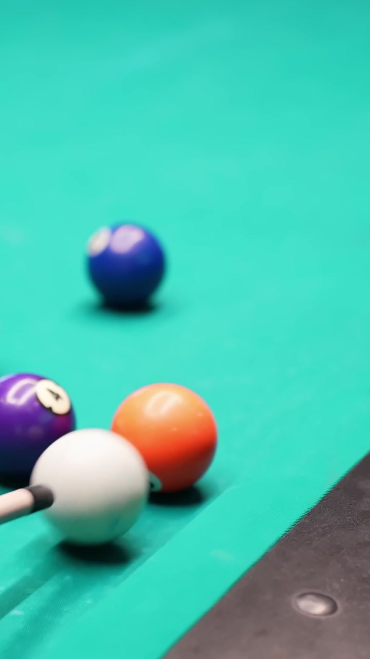 Vertical close-up of cue ball beside a cluster of numbered billiards spheres on teal felt, shallow depth of field and soft motion blur for indoor pool game b-roll.