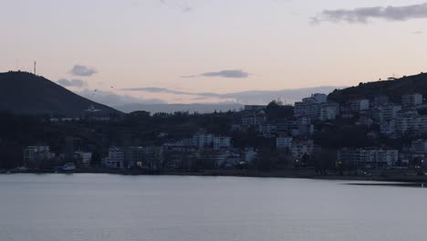 Eine-Statische-Aufnahme-Eines-Verträumten-Seestrandes,-Waldberge-Im-Hintergrund,-Europäische-Sonnenuntergangsdämmerung,-Fliegende-Vögel,-Blaues-Klares-Wasser,-Stabiles-4K-Video