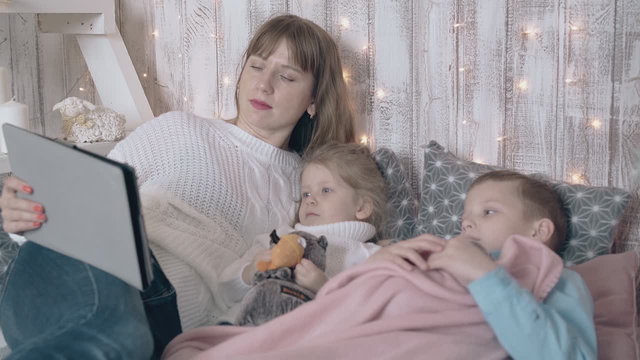 los niños ven dibujos animados en la tableta gris acostados en la cama con la madre