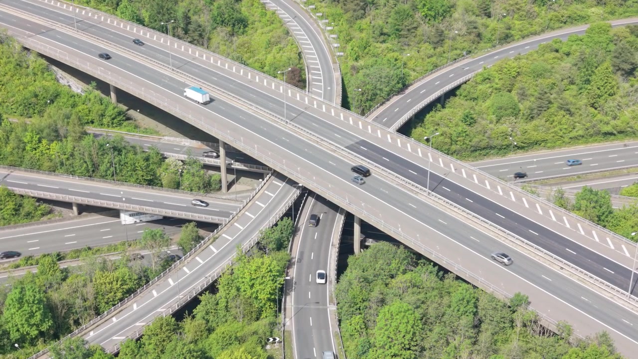 Aerial view of a complex multi-level highway interchange. Cars, trucks, and buses drive on interwoven roads, showing active traffic flow. Green trees surround the vast infrastructure during daylight.