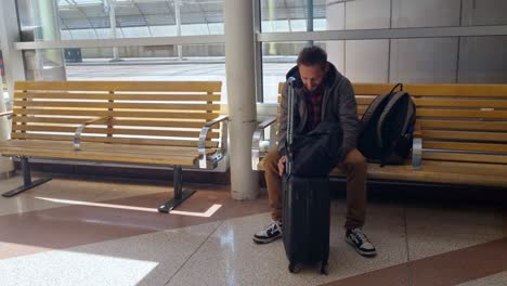 a man waits at a bus stop, visibly bored, spinning his suitcase in slow circles to pass the time. a quiet, everyday moment of travel and impatience
