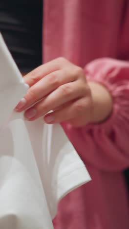 vista inferior del cuerpo de una dama que lleva una camiseta rosa sin botones evaluando pensativamente una manga de camiseta de polo blanca con escritura débil en ella, con pantalones de vaqueros cuidadosamente dispuestos en un estante de ropa detrás
