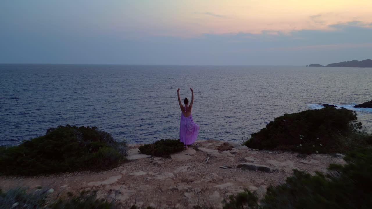 Woman meditating in yoga position on a cliff overlooking the sea at sunset in Ibiza. Smooth aerial view flight overflight flyover drone