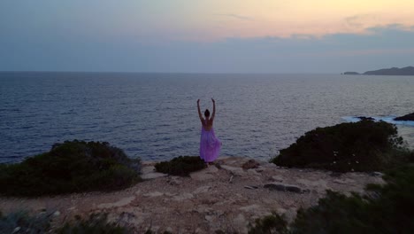 woman meditating in yoga position on a cliff overlooking the sea at sunset in ibiza. smooth aerial view flight overflight flyover drone