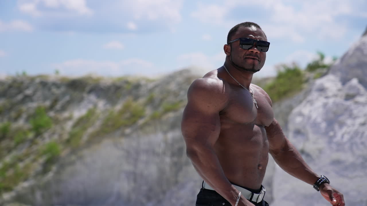 Waist up portrait view of the athletic naked man wearing glasses walking outside and showing his naked torso. Man posing at the sandy beach
