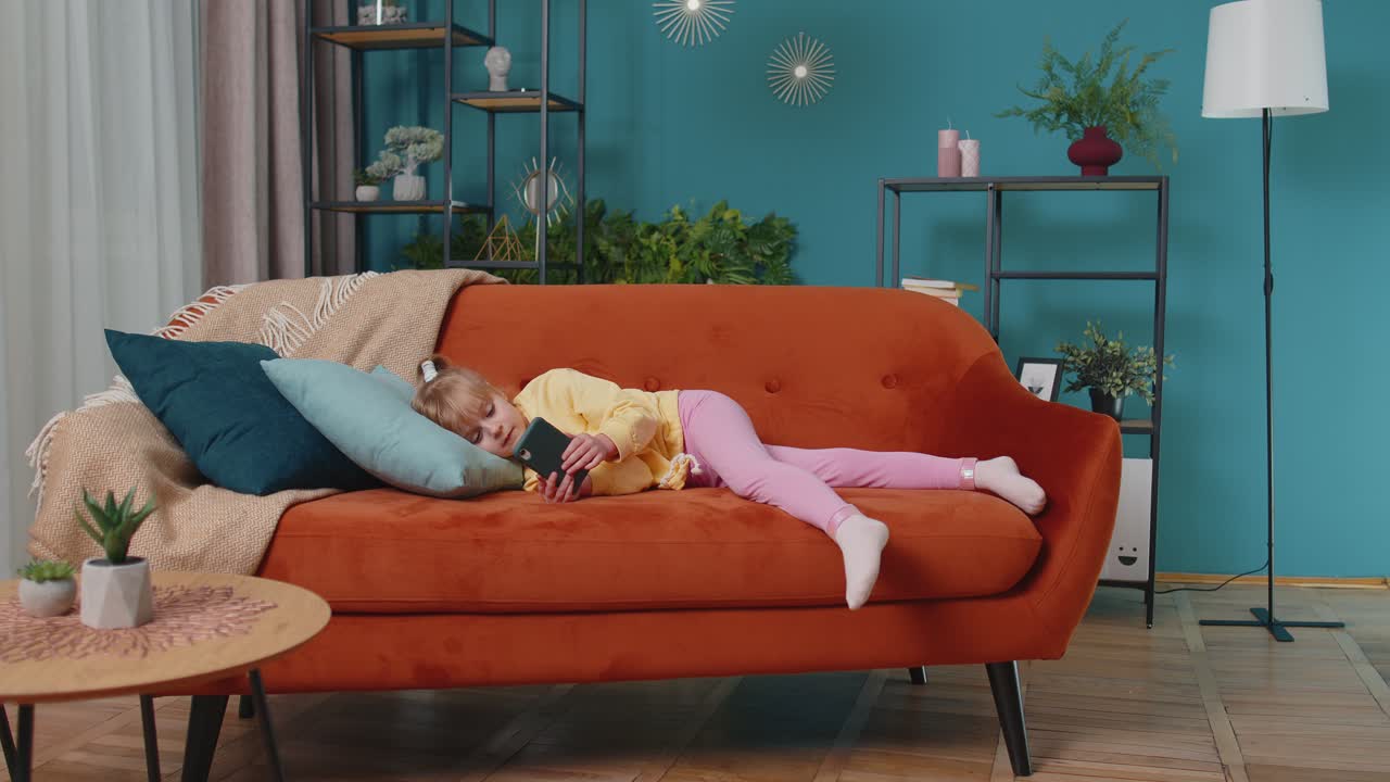 Child toddler hold smartphone watching online cartoons chatting with friends on sofa alone at home