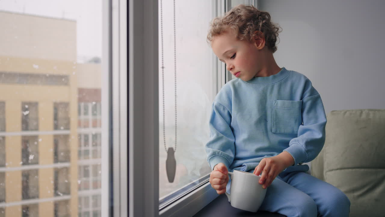 baby boy with blond curly hair is sitting alone in apartment and watching snow in winter day cute toddler