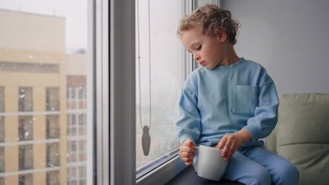 baby boy with blond curly hair is sitting alone in apartment and watching snow in winter day cute toddler