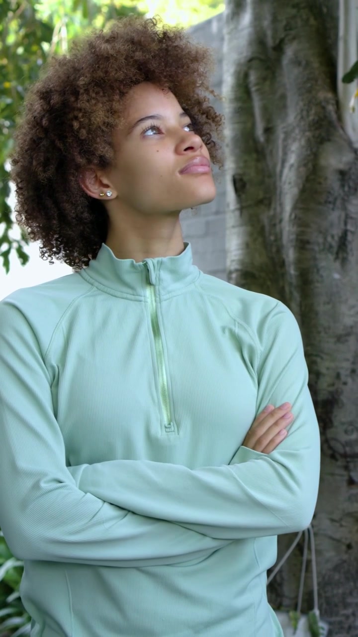 Video verticale: Donna con top color menta che alza lo sguardo verso la luce tra il fogliame in giardino, vicino a un tronco d'albero.
