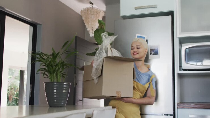 Donna felice di etnia mista che cammina con una scatola con pianta a casa al rallentatore