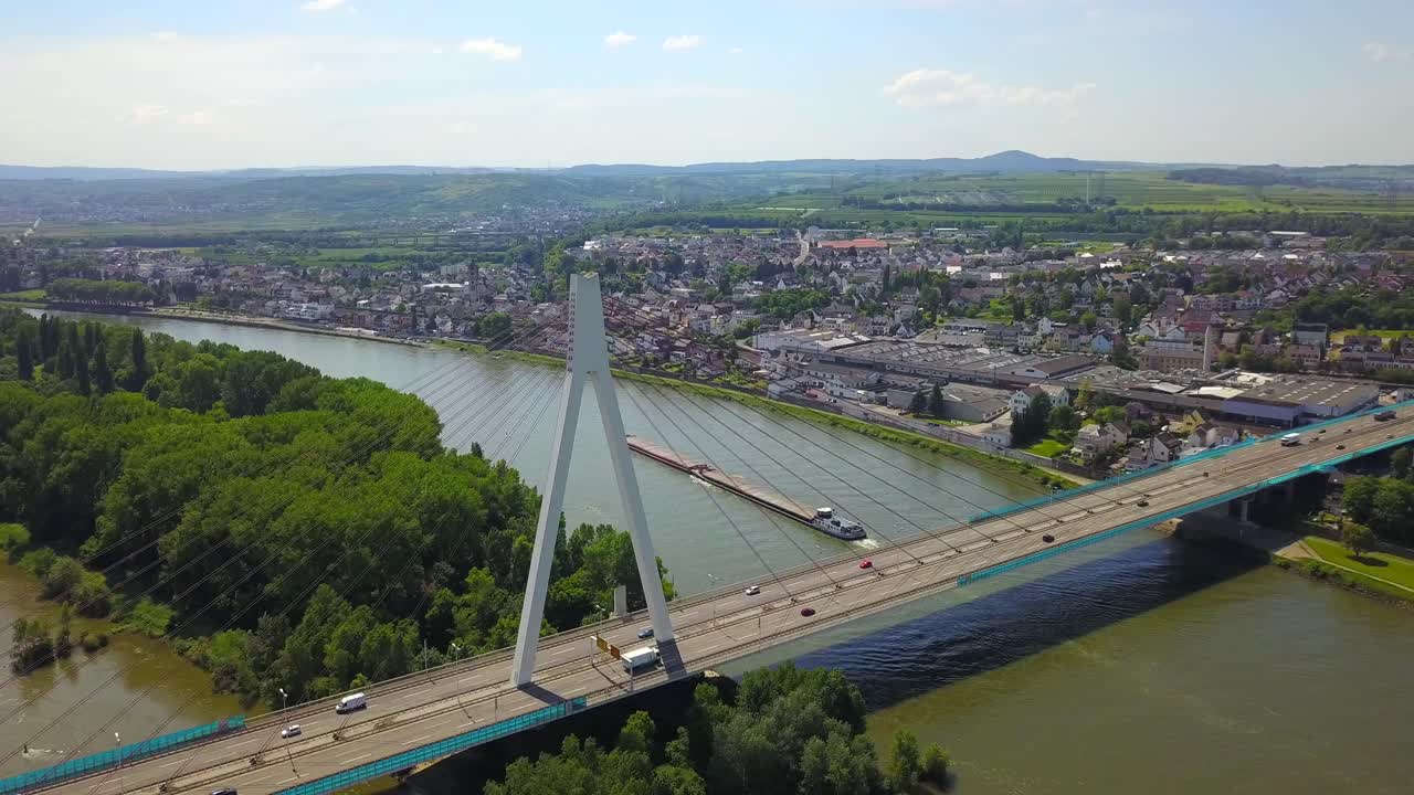 Stunning drone perspective of the Neuwied Rhine Bridge, where vehicles flow along the highway while cargo ships travel beneath on the river. The transportation hub connects commerce and daily life.