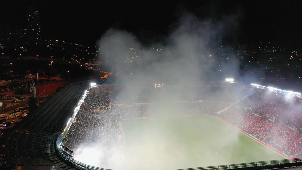 Denso fumo pirotecnico che riempie lo Stadio Teddy durante una partita di calcio, circondato da tifosi esultanti di notte.