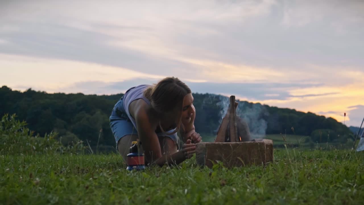 Woman kneeling on grass blowing on small campfire to ignite flame near lake at sunset, capturing outdoor survival action and camping activity in Savci Slovenia