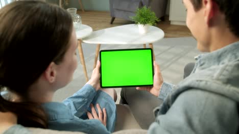 over the shoulder view young couple at home sitting on a sofa using with green mock-up screen tablet. man and woman using touchscreen device, browsing internet. concept of green screen and chroma key