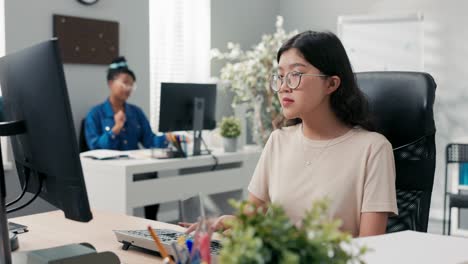 office workers are sitting at desks in front of computers fooling around woman with korean beauty is working diligently another girl disturbs by throwing paper airplane at her colleague returns favor