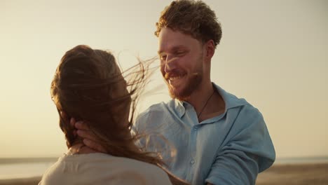 A-bearded-man-in-a-blue-shirt-fixes-his-girlfriend's-hair-in-a-windy-weather-outside.-A-romantic-meeting-against-the-yellow-sunset